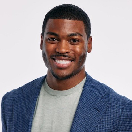 A smiling man with short, neatly trimmed hair and a well-groomed beard, wearing a light gray shirt and a blue checkered blazer, stands against a plain white background. His friendly and approachable demeanor is highlighted by his broad smile and confident posture.