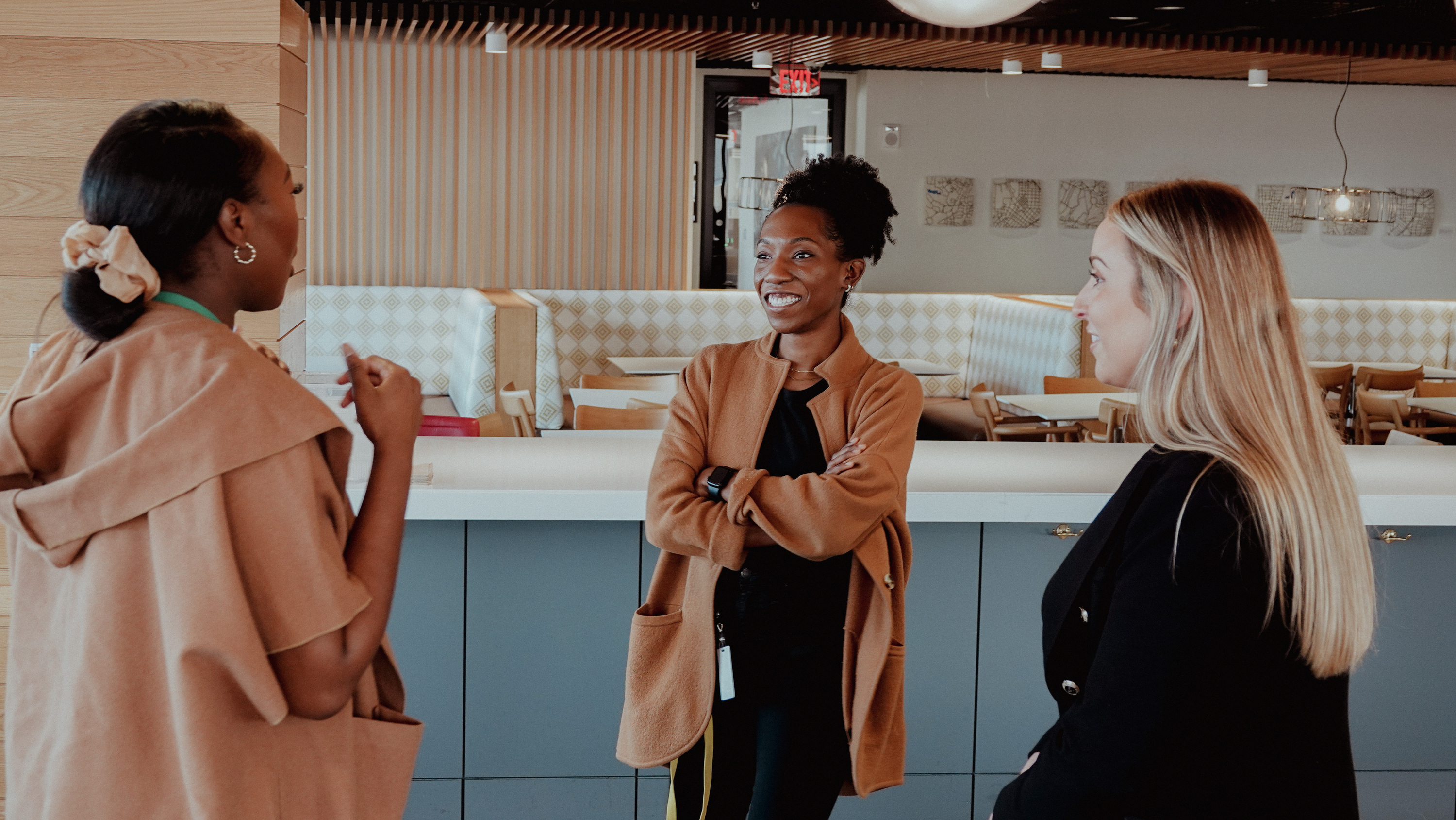 Three individuals engaging in a friendly conversation in a modern, well-lit communal space with wooden accents and contemporary furniture in the background.