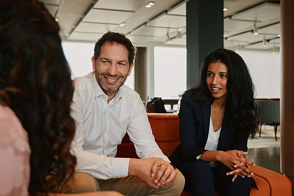 A man and women sit on a couch in an office setting. They are speaking to a woman slightly out of frame who is sitting, facing them. 