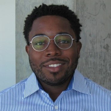 Headshot of a man with short curly hair, wearing glasses and a blue and white striped shirt, smiling against a light background with a textured wall.