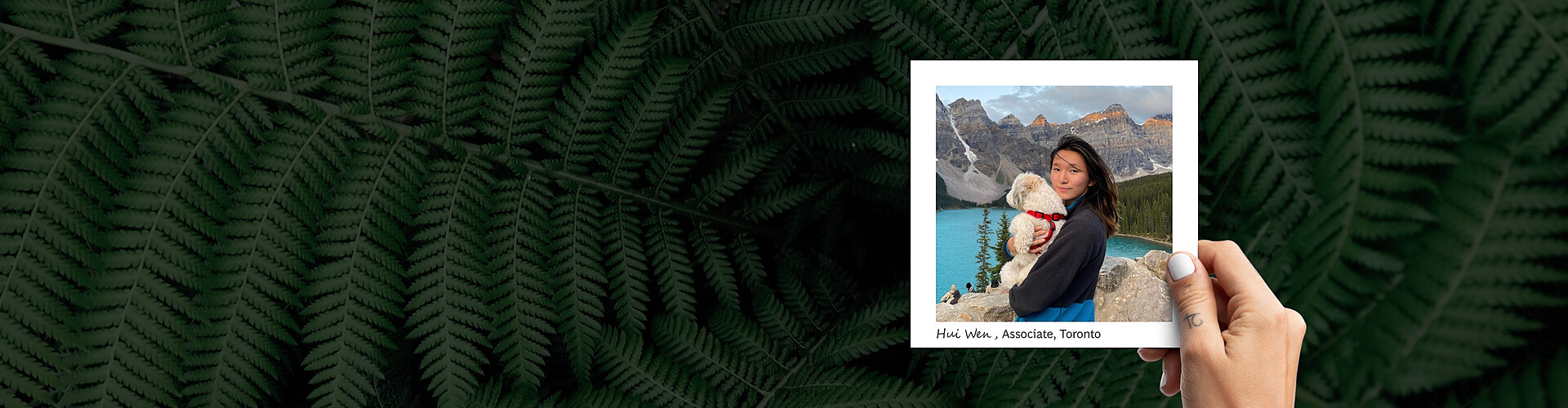 A woman's hand with a thumb tattoo and white nail polish holds a white frame. In the frame, a woman is holding a small white dog standing near the edge of a cliff. Behind her is a large blue lake, a mountain range, and a forest of pine trees. In the background behind the frame is a zoomed in look at fern leaves.