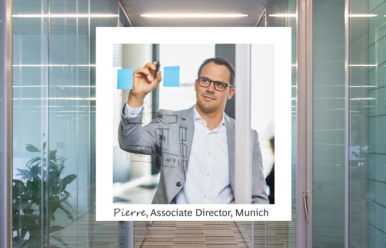 The image features Pierre, an Associate Director in Munich, actively engaged in a strategic planning session within a modern office environment. He is framed within a white border, standing and writing on a glass wall with a marker, demonstrating a moment of thought or presentation. Pierre is dressed smartly in a tailored grey suit and glasses, which complements the professional setting characterized by sleek, glass partitions and soft indoor lighting.