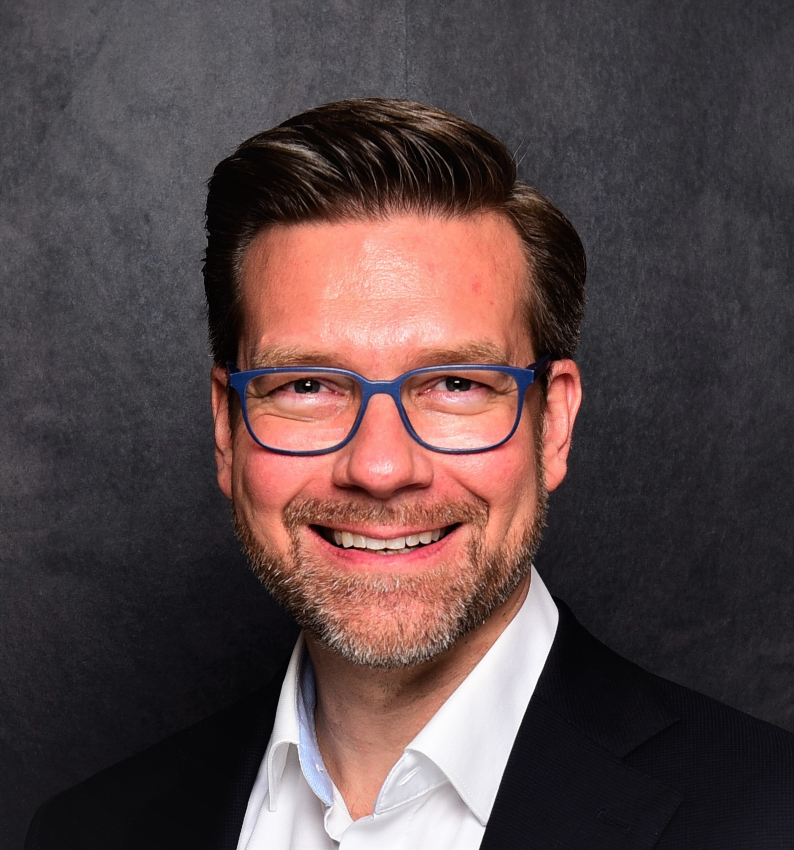 Headshot of a man with neatly styled dark hair, wearing blue-framed glasses, a white shirt, and a dark suit jacket, smiling warmly against a dark textured background.