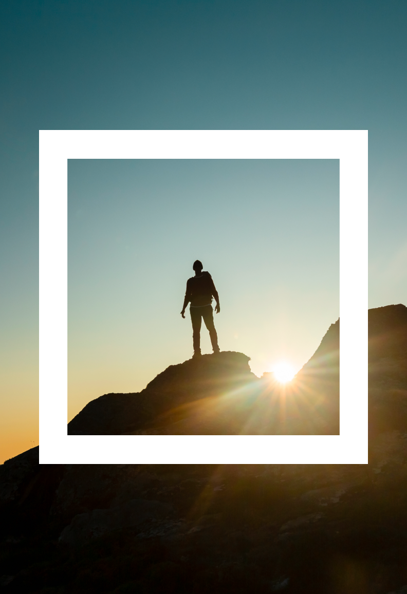 Persona in piedi su una roccia al tramonto, in controluce, con il sole che sorge alle spalle.