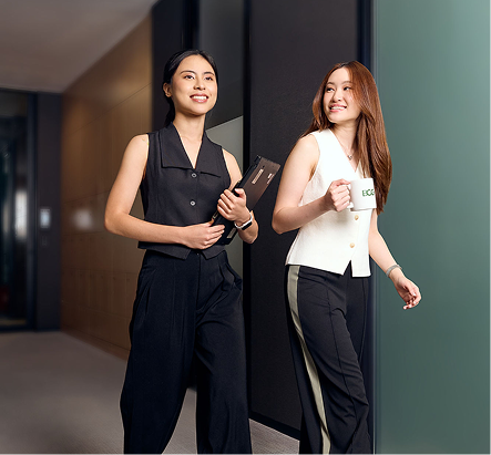 Two women walk in an office hallway. One, wearing a black top and black pants, carries a closed laptop. The other, wearing black pants and a white top, carries a BCG mug. They are both smiling.