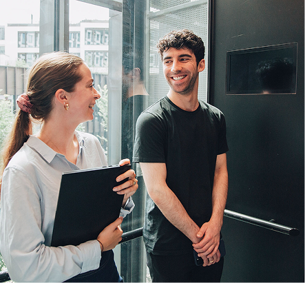 A man and a women are in an office building, standing in front of a glass window. The women is holding a laptop. They are smiling at one another.