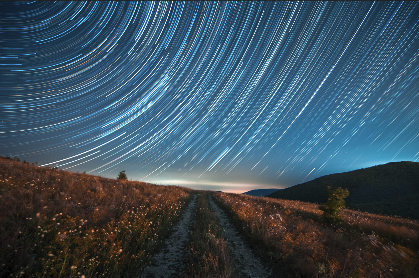 Sentiero tra i campi sotto un cielo notturno con scie luminose di stelle in movimento.