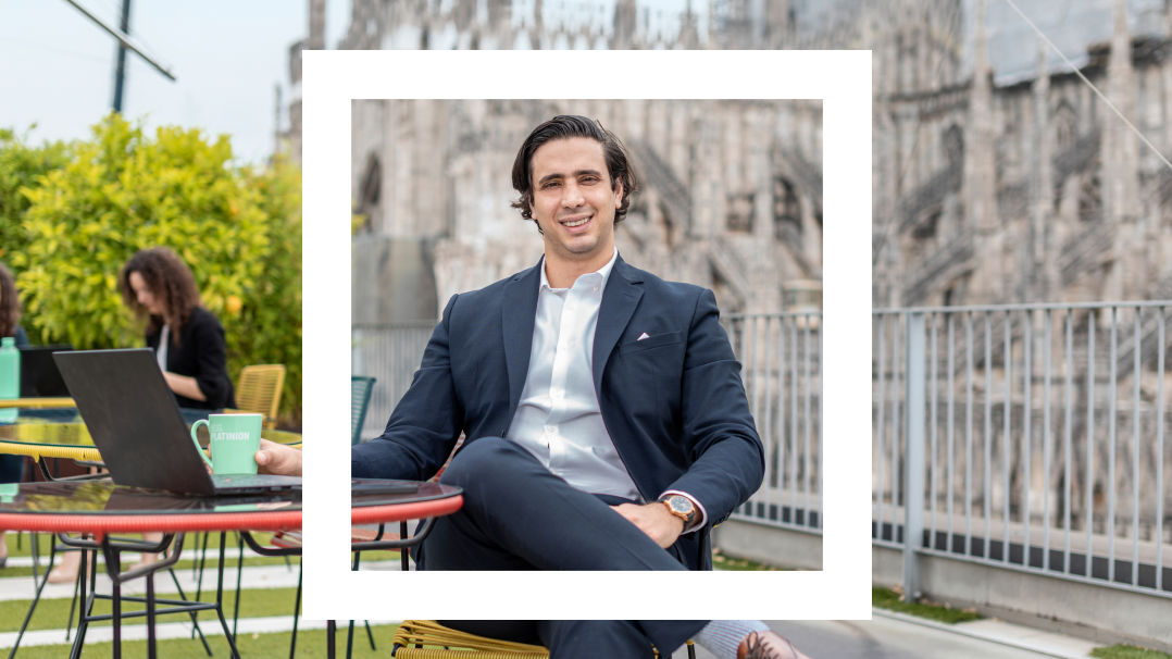 The image depicts a professional man, seated confidently at a café table outdoors, framed within a white border. He is dressed in a sharp navy suit with a white shirt and is smiling. In the background, other individuals can be seen working at similar tables, set against a lush, urban park and the intricate architecture of a large, historic cathedral. 