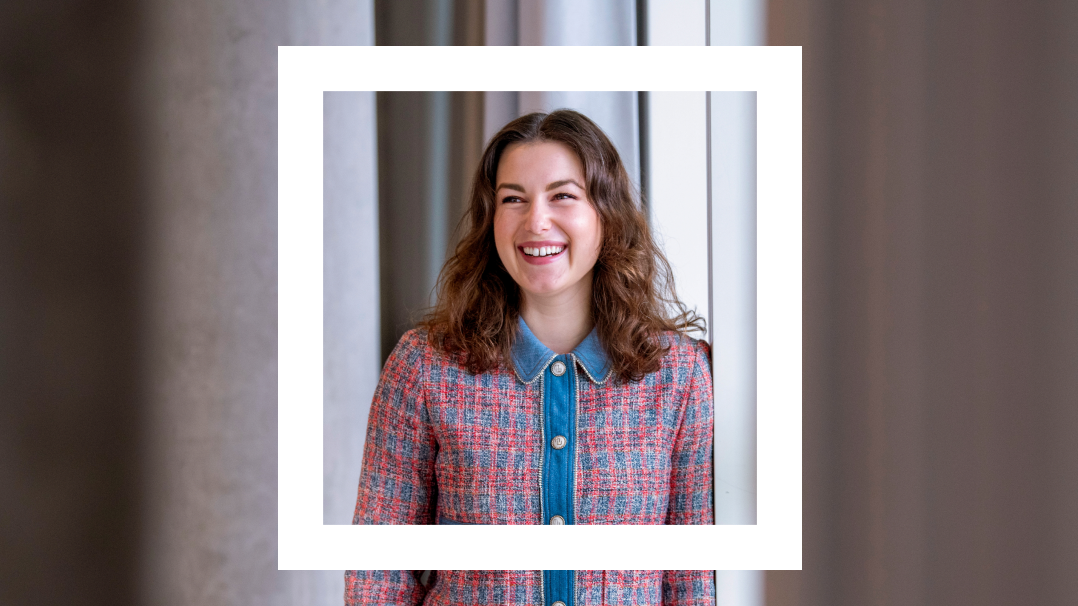 The image showcases a young woman, captured in mid-laughter, framed within a white border. She is dressed in a stylish tweed jacket with a denim collar, embodying a blend of professional and casual style. The backdrop suggests an office environment with soft lighting and neutral colors, enhancing the warmth and friendliness evident in her expression. 