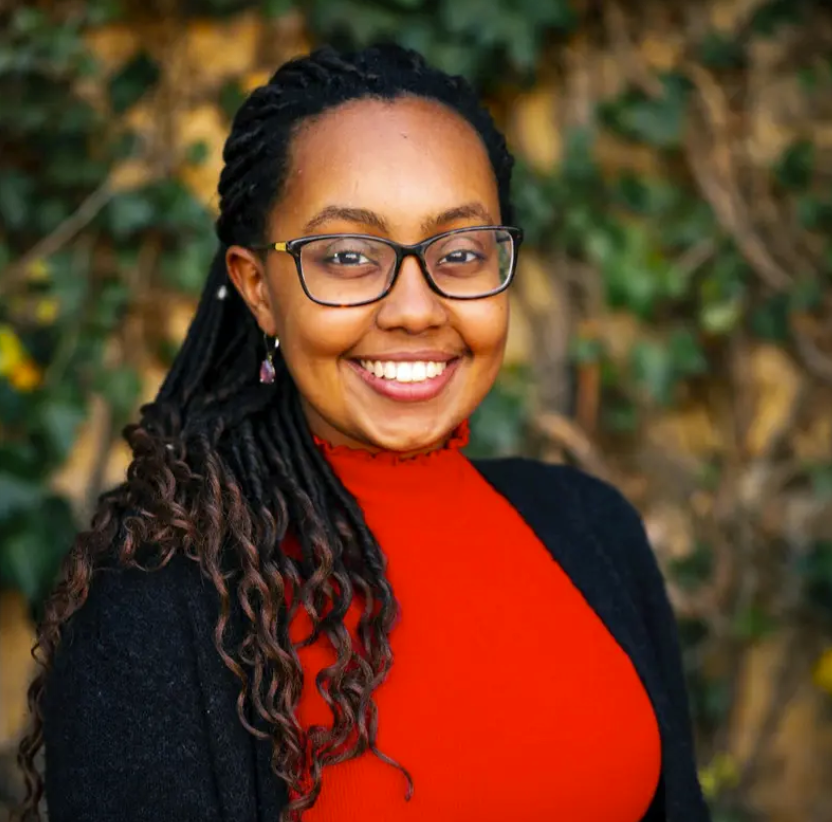 A smiling woman with glasses and long, dark braided hair, wearing a red top and black cardigan, stands in front of a background of green ivy leaves. She exudes confidence and warmth, with a natural and friendly demeanor. The ivy backdrop provides a contrasting natural element to her vibrant outfit.