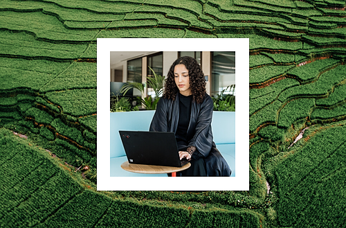 A woman with curly hair, dressed in dark clothing, working on a laptop while seated in a modern indoor space, framed within an overlay of lush green terraced fields in the background.