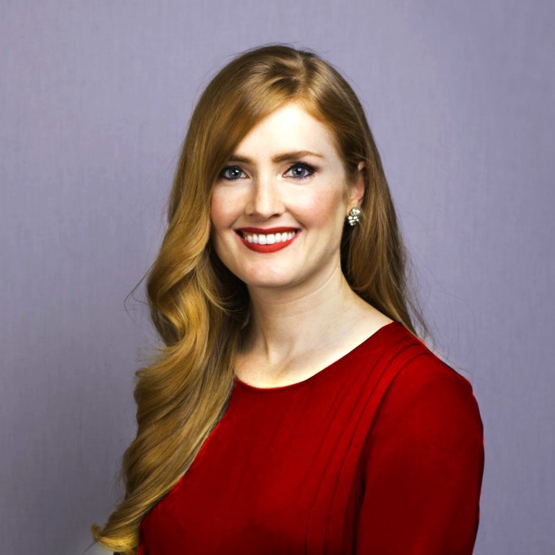 Portrait of a woman with long red hair, wearing a red dress and silver earrings, smiling warmly against a soft purple background.