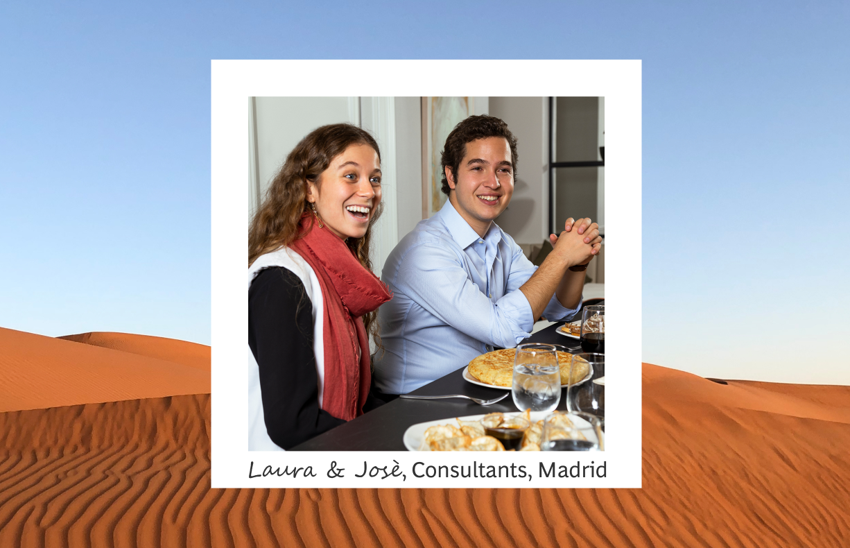 The image shows Laura and José, consultants in Madrid, sharing a joyful moment at a dinner table, framed against a surreal background of desert dunes. They are both dressed in casual office attire, with Laura wearing a scarf, enhancing the informal yet professional vibe of the meeting. The dining setting includes glasses and plates, suggesting a relaxed business dinner. 