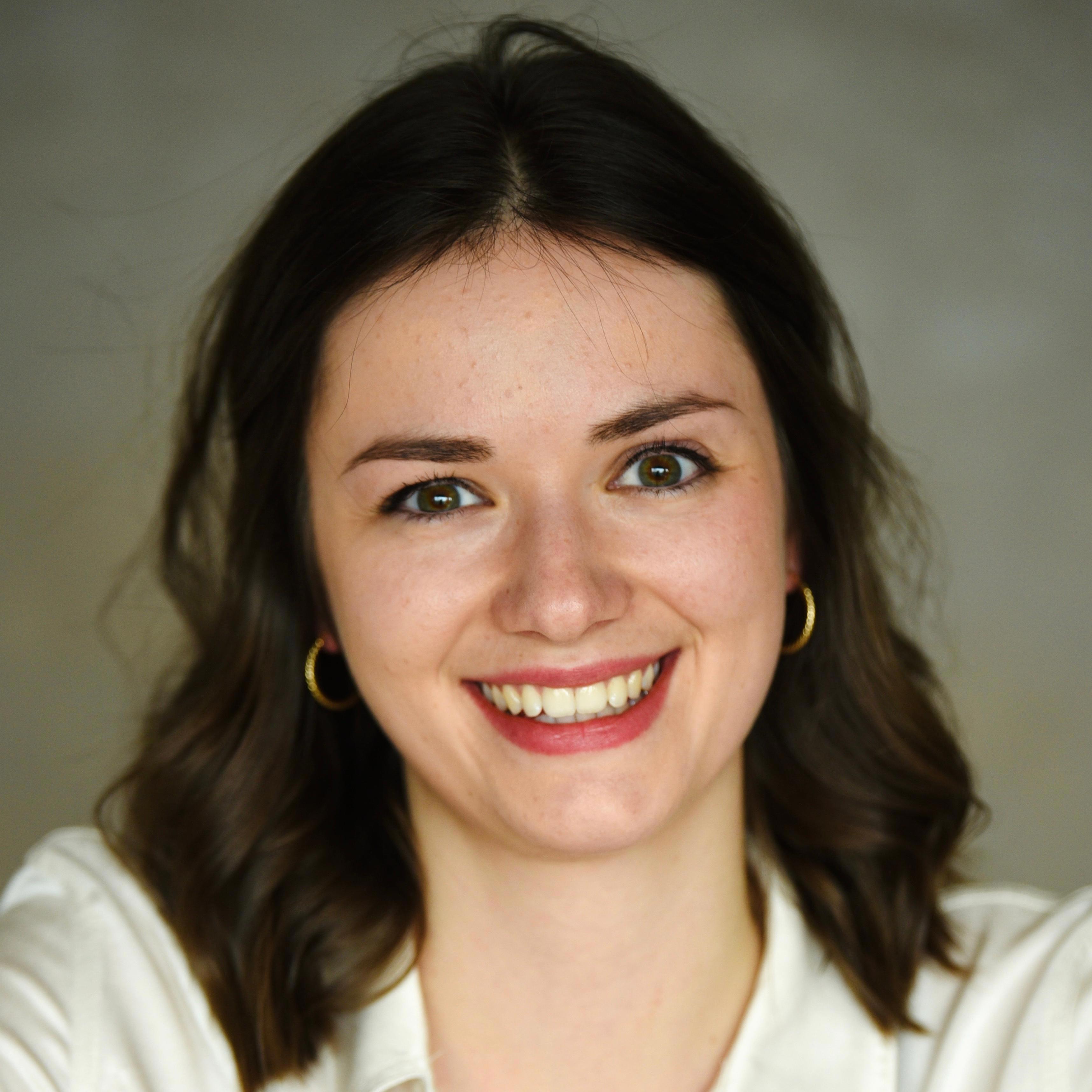 A woman with shoulder length brown hair wearing a white button down smiles at the camera in front of a grey background.