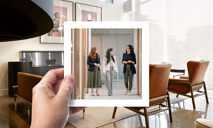 A hand is holding a framed photo of three women walking in a office hallway. They are walking toward the camera and appear to be in conversation with one another. The framed photo is situated over a background image of an office with brown leather chairs and framed artwork on the wall.