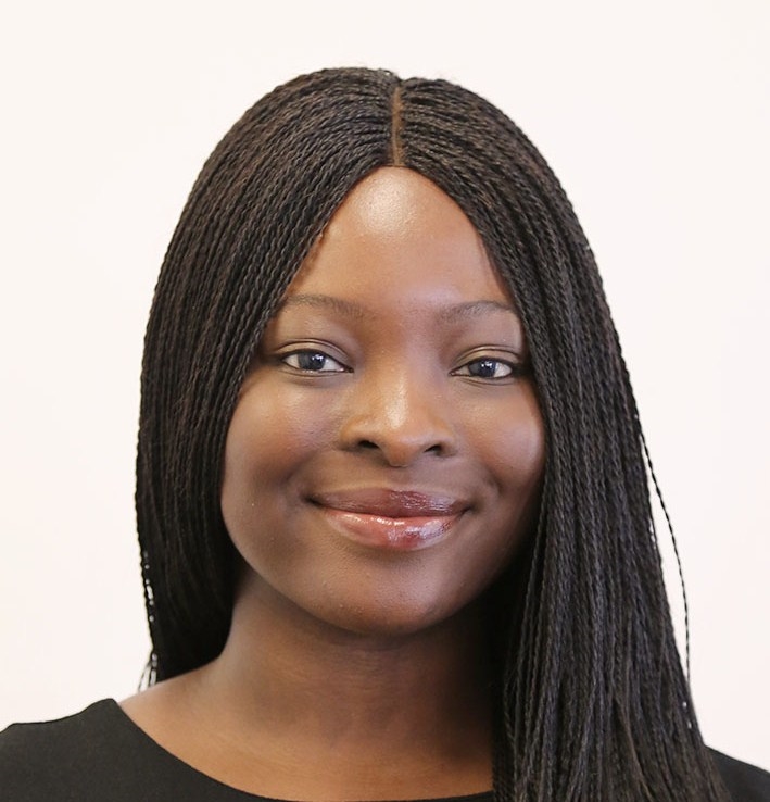 A smiling woman with long, neatly braided hair, wearing a black top, stands against a plain white background. Her friendly and serene expression, along with her clear complexion and glossy lips, exudes confidence and warmth.