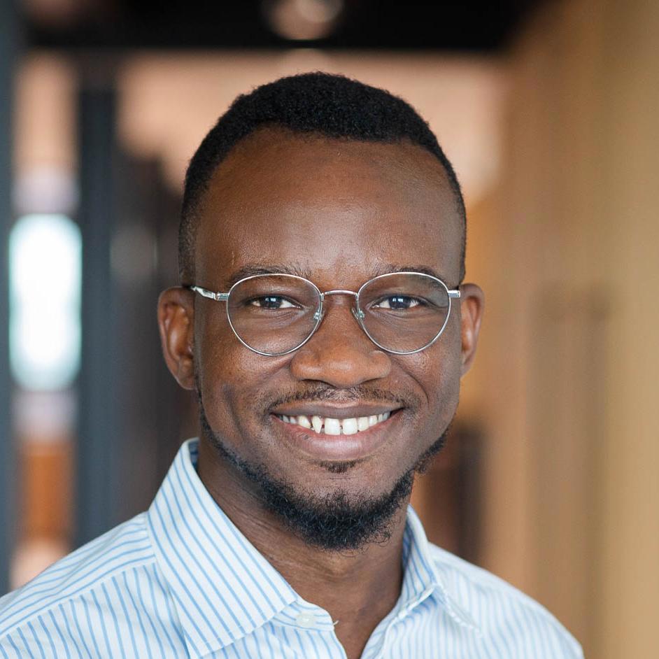  A smiling man with short hair and a well-groomed beard, wearing round glasses and a light blue and white striped shirt, stands indoors with a softly blurred background. His friendly and approachable demeanor is highlighted by his warm smile and clear, bright eyes.