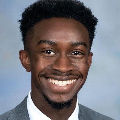 Portrait of a smiling individual wearing a suit and tie, with short curly hair and a friendly demeanor against a neutral background.