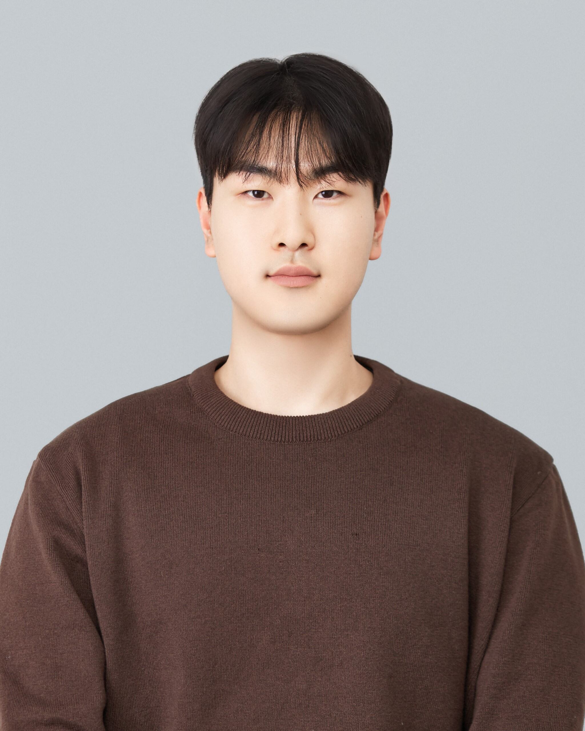 Portrait of a young man with short black hair, wearing a brown sweater, against a plain light gray background.