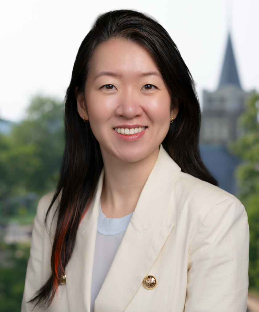 The image shows a professional woman with long black hair, wearing a cream-colored blazer with gold buttons. She is smiling, and the background features greenery and a blurred building with a pointed roof.