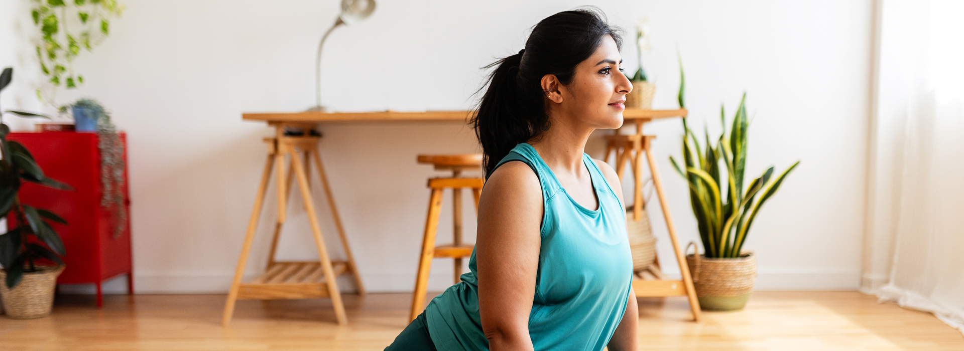  Person practicing yoga on a mat in a bright indoor space with wooden floors, stools, and plants visible, suggesting a calm and wellness-focused environment.