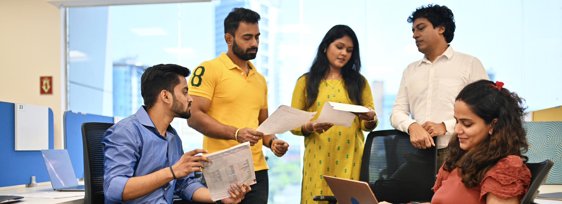 Group of people gathered in a modern office workspace, reviewing printed documents and talking together near desks and office chairs. Laptops and office equipment are visible, suggesting a collaborative learning environment.