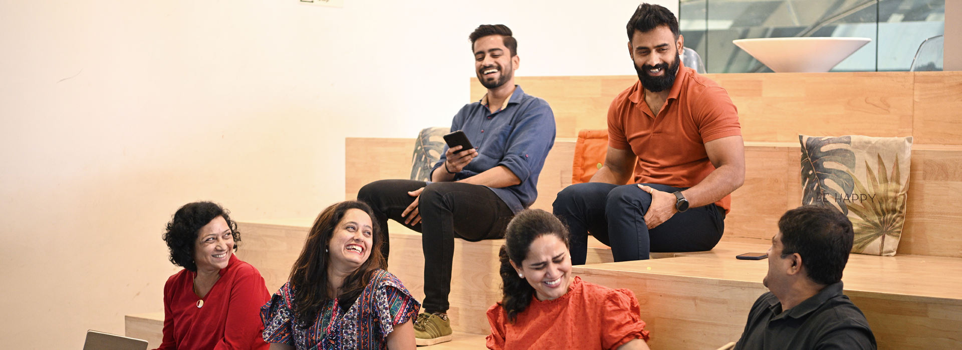 Group of people seated on tiered wooden steps in an open indoor space, talking together in a casual setting. Cushions and light-colored walls are visible, creating an informal meeting environment.