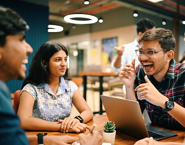  People seated at a table in a modern workspace, engaged in discussion while reviewing a laptop. The setting includes overhead lighting, wood surfaces, and a collaborative, informal layout.