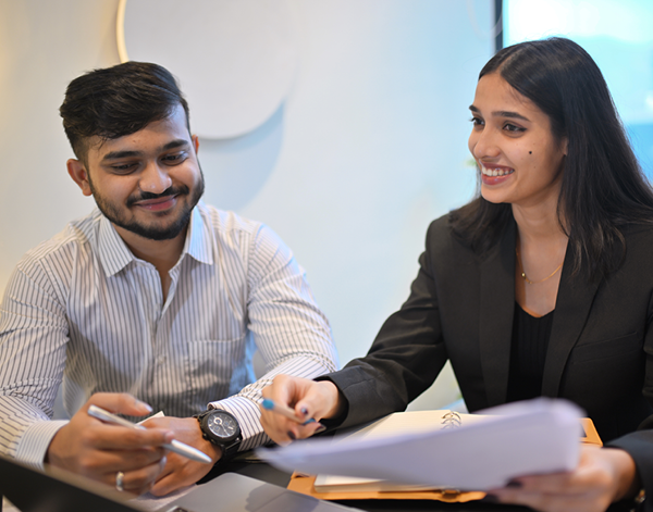 Two people seated at a desk reviewing printed documents together, with one person holding papers and the other gesturing while speaking. The setting appears to be a formal office workspace.