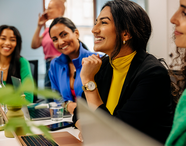 Group of people seated at a table with laptops open, participating in a discussion in a shared office space. Notebooks, greenery, and a bright, modern work environment are visible.