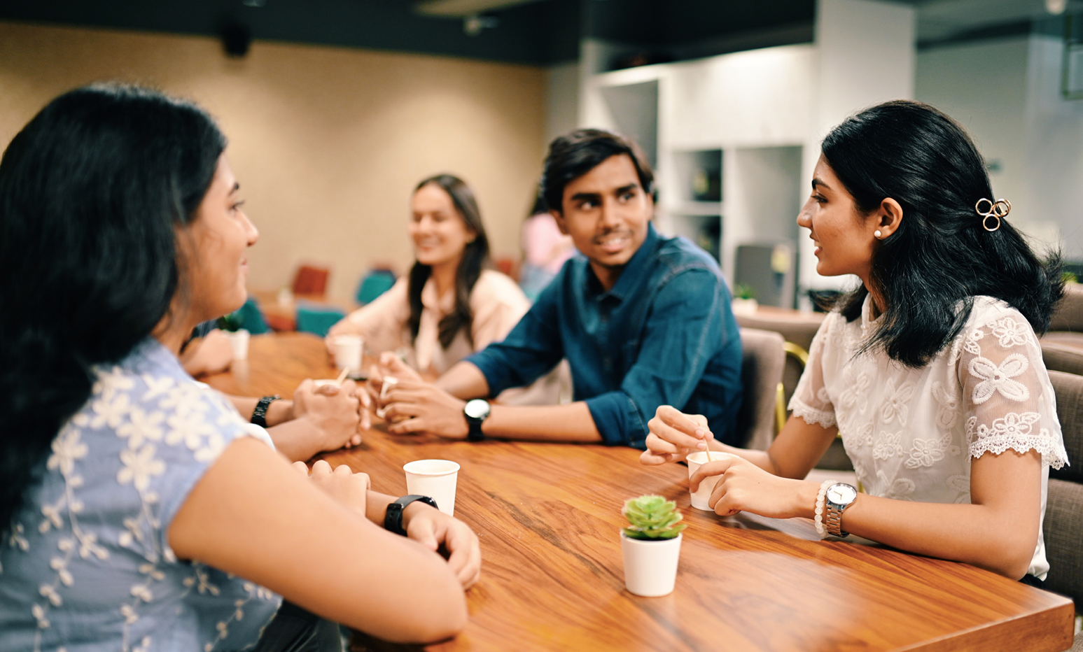 Group of people seated around a wooden table in a modern office, participating in a small group discussion. Coffee cups and a small plant are on the table, with soft lighting and office shelving visible in the background.