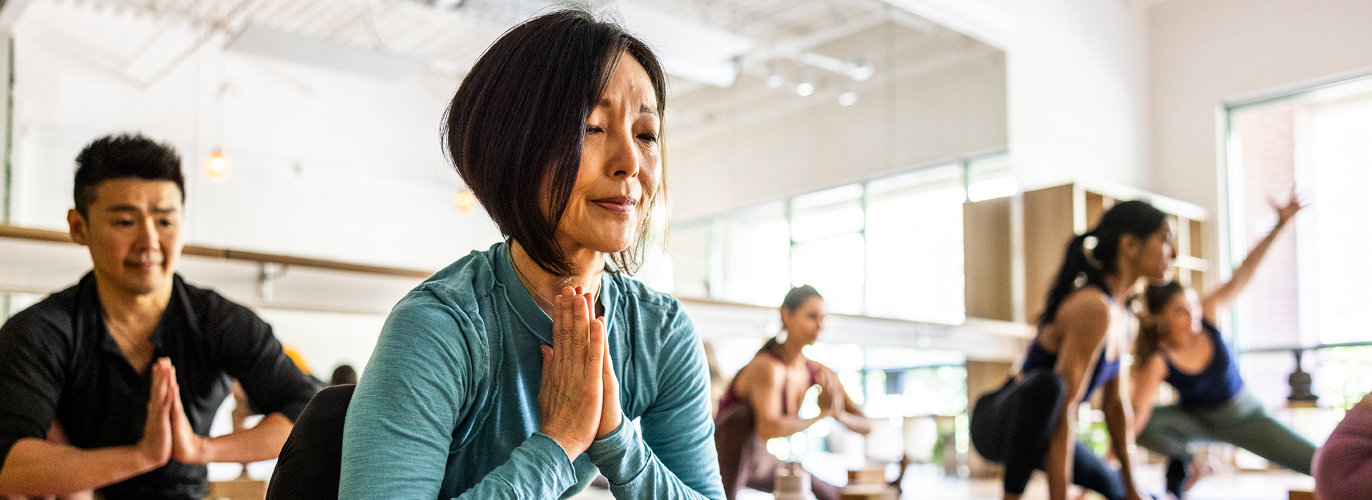 Group of people participating in a yoga class inside a bright studio, with several individuals seated on mats in a prayer pose while others stretch in the background. Natural light fills the space, and mirrors and windows are visible.