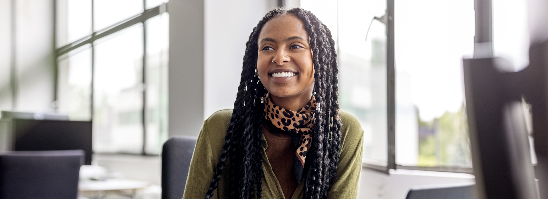 Person seated in a modern office environment with large windows, wearing a patterned scarf and a green top, with office chairs visible in the background and natural light filling the space.