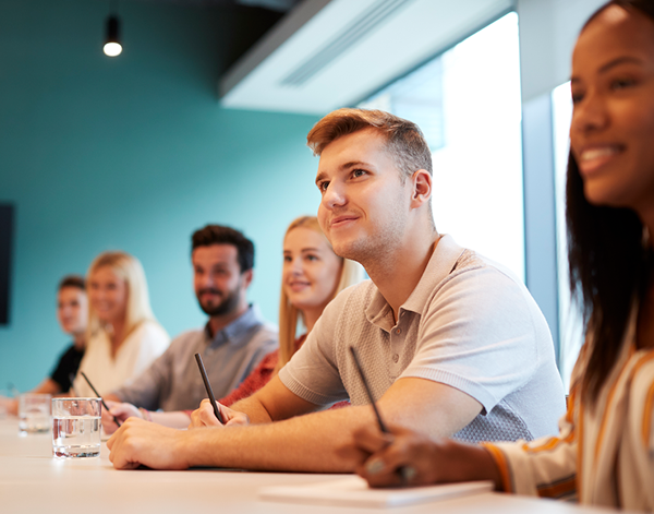  Several people seated at a conference table in a modern meeting room, taking notes during a discussion. The space features teal-colored walls, overhead lighting, and notebooks and glasses of water placed on the table.