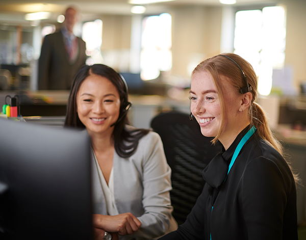  Two people seated at a desk in an open office environment, looking at a computer monitor together. Office workstations and a colleague standing in the background are visible, with natural light coming through windows.