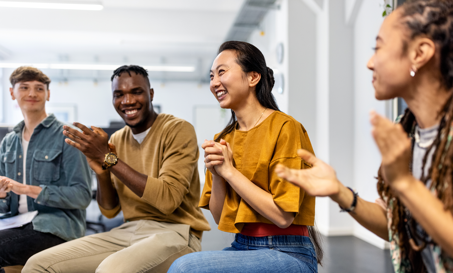  Group of people seated side by side in a bright indoor space, clapping during a group activity or discussion. Casual clothing is worn, and the setting features white walls, overhead lighting, and an open, modern layout.