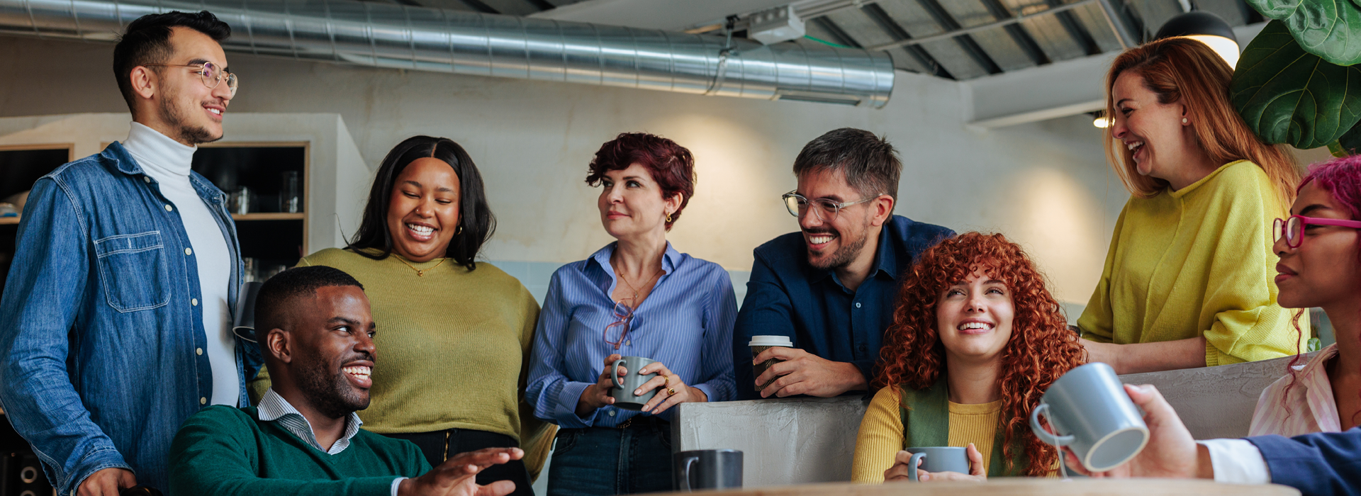 Group of people gathered around a counter in a casual office setting, standing close together and engaged in conversation. Mugs, a countertop, and overhead lighting are visible in the space.