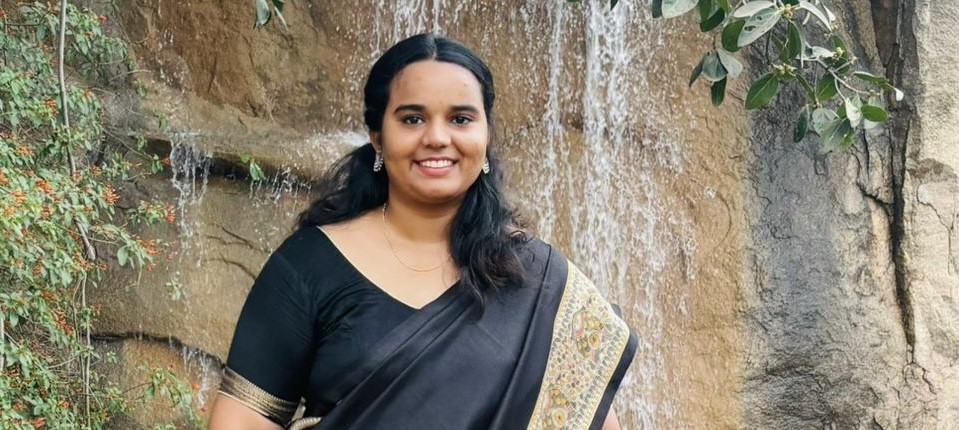 Person standing outdoors near a small waterfall and rock formation, wearing a dark traditional outfit with a patterned border. Green plants and flowing water create a natural, serene background.