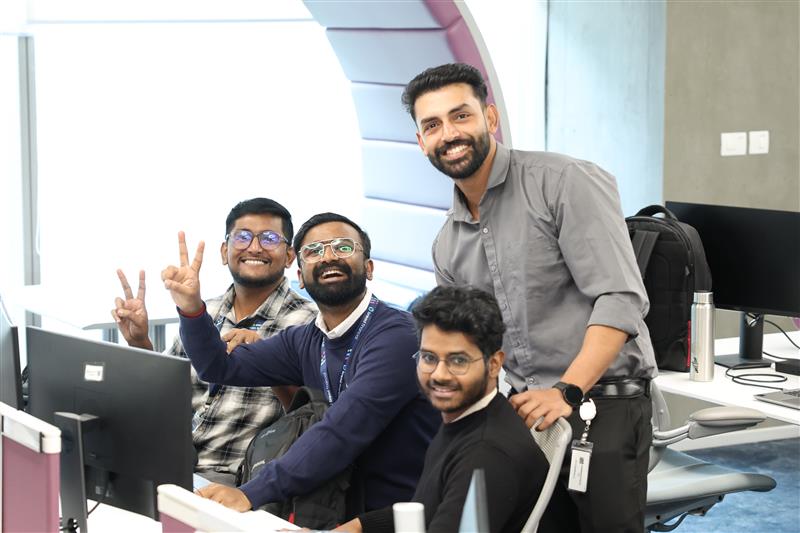 Group of associates gathered around desks in an open office, collaborating while looking at computer screens. Office chairs, monitors, and a bright workspace layout are visible.