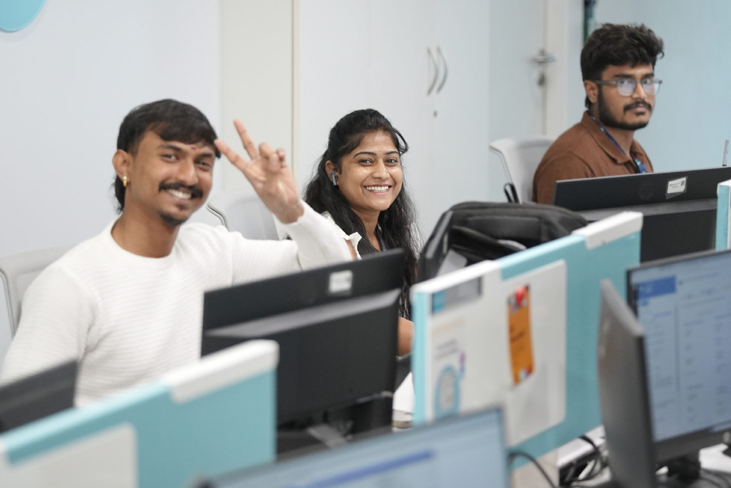 Associates seated at workstations in an open office, using desktop computers and participating in conversation. Desk dividers, monitors, and overhead lighting are visible.