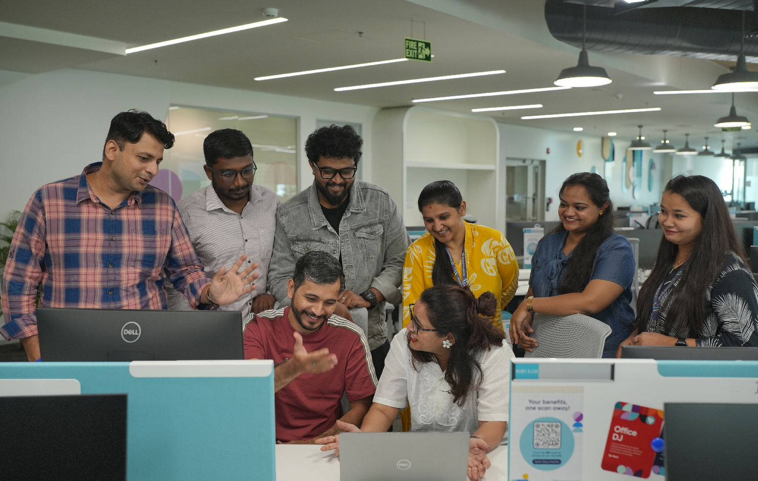  Group of people collaborating around desks in a modern open office workspace, discussing work while viewing laptops. The space features teal desk partitions, overhead lighting, and an open, contemporary layout with multiple workstations.