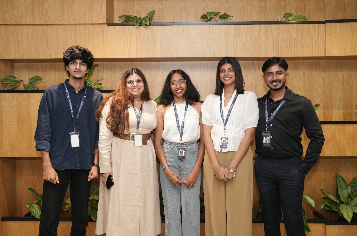 Group of associates standing side by side in an indoor office area, wearing work badges. Wooden wall panels, shelving, and potted plants are visible behind them.