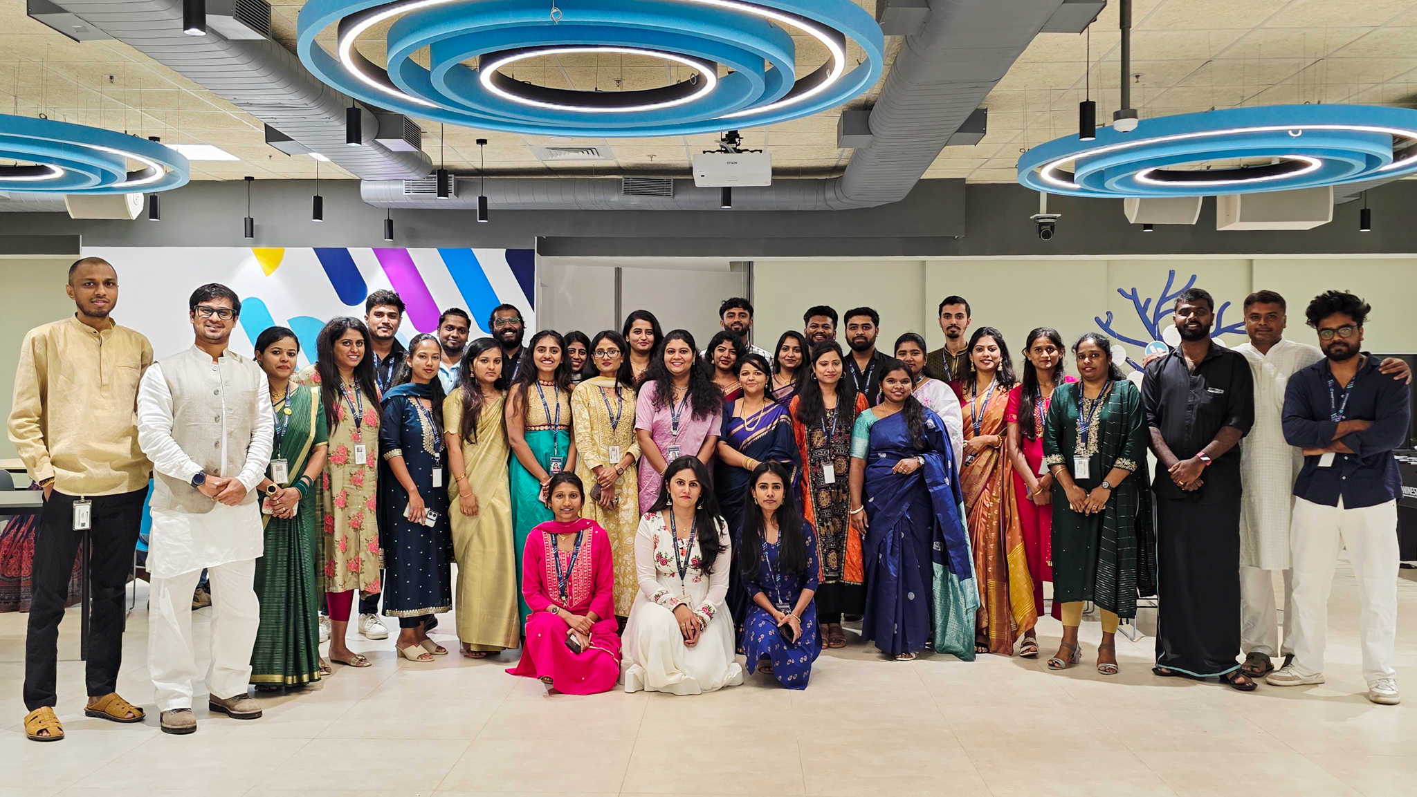  Large group of associates standing together in a modern office space, wearing a mix of traditional and contemporary clothing. Circular ceiling lights, open floor space, and a colorful wall design are visible in the background.
