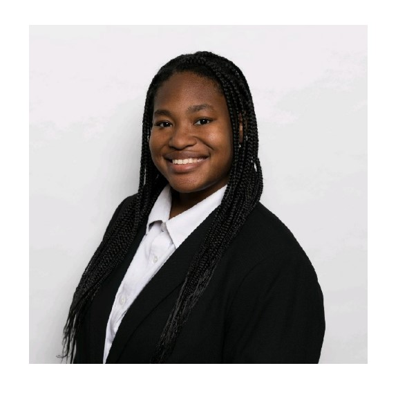  Person standing against a plain light-colored background, wearing a white collared shirt and a dark jacket, with long braided hair draped over the shoulders.