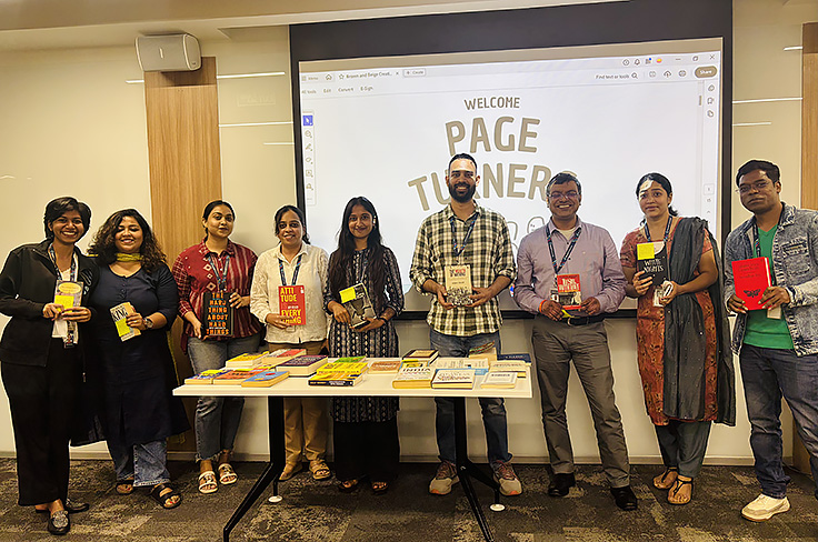  Group of associates standing behind a table with books and materials displayed in an indoor event space. A presentation screen and wall signage are visible in the background.