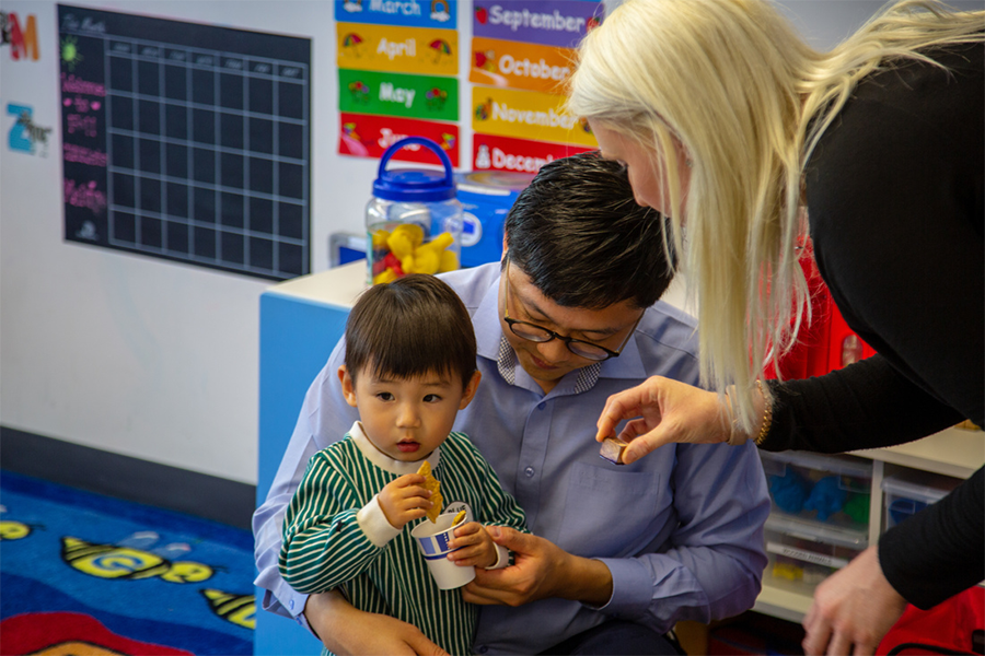 Child eating a snack with two adults nearby in a classroom.