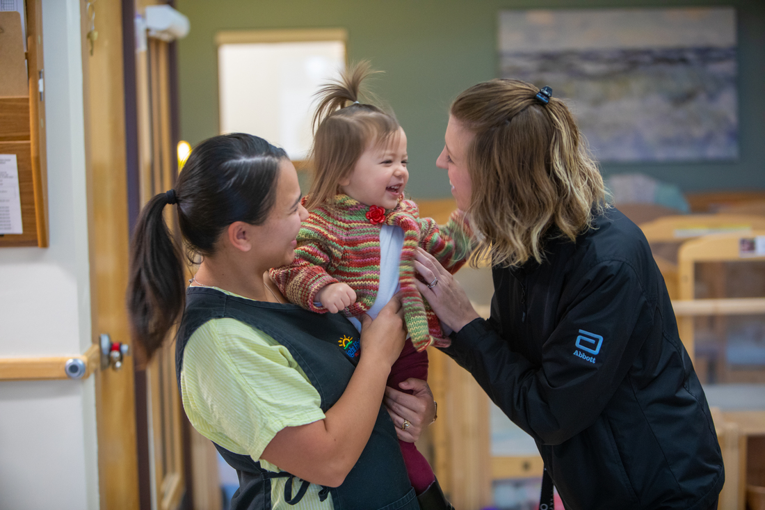 A teacher and a parent interacting with a young child inside a bright, organized classroom setting.