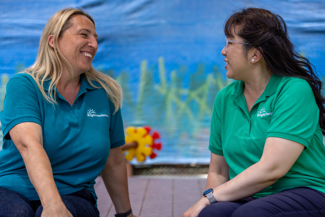 Two Bright Horizons educators engaged in conversation in a colorful child care setting, with a gear toy and aquatic-themed mural in the background