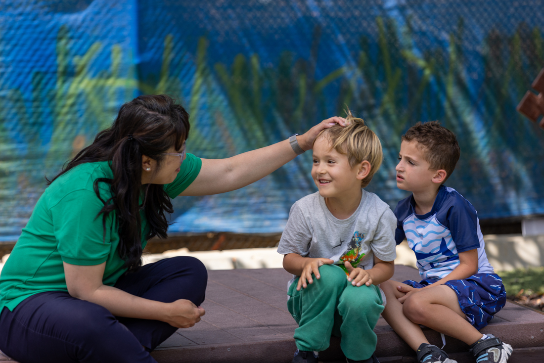 An educator sitting outdoors gently interacting with two young children during playtime.