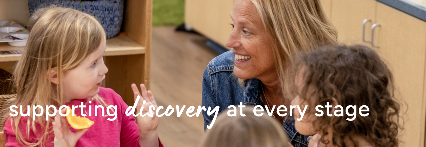 A woman engages with three young children in a classroom, one child holding an orange slice.